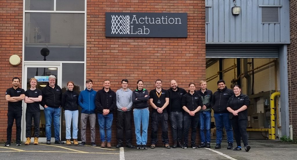 Fourteen people in Actuation Lab team outside in front of building with Actuation Lab logo
